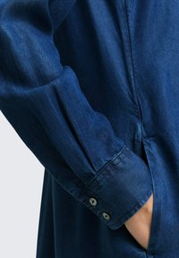 Chemise en denim de couleur bleu foncé, avec des manches longues, des poignets à boutons et une poche latérale, présentant une texture lisse.