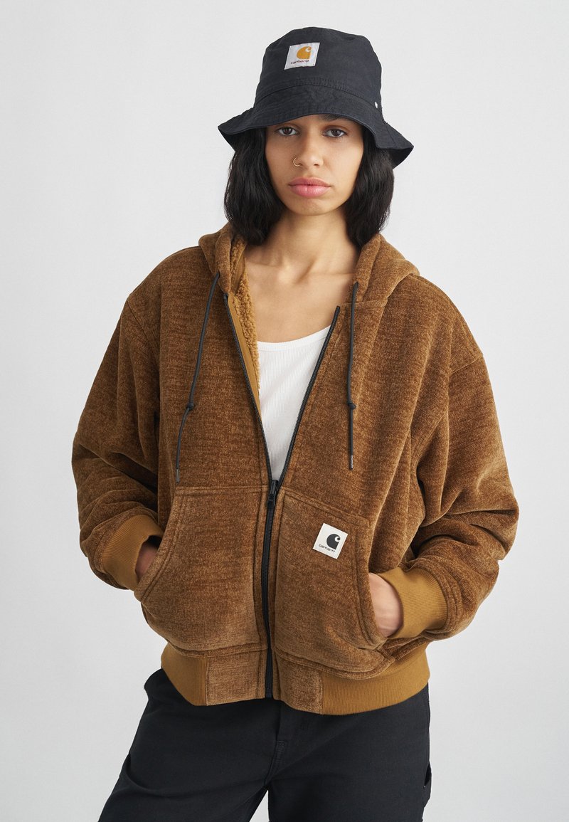 Jovem vestido com um hoodie de fleece castanho com fecho, chapéu preto tipo balde, camisa branca e calças pretas, de pé com as mãos nos bolsos.