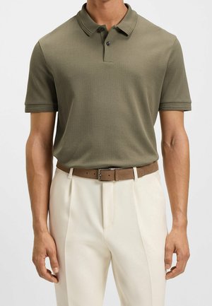 Homme portant un polo vert olive à manches courtes, rentré dans un pantalon blanc cassé avec une ceinture marron clair, debout devant un fond uni.