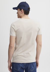 Beige pamut póló egyszerű dizájnnal és rövid ujjakkal, kék farmernadrággal és sötétkék sapkával viselve. Nincsenek látható minták.