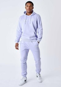 Sudadera de color púrpura claro y joggers a juego, con un bolsillo frontal, capucha con cordón y logo bordado. Zapatillas blancas completan el conjunto.