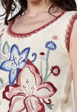 Haut sans manches en maille crème avec grandes fleurs roses et bleues brodées et tiges vertes, porté par une personne aux cheveux bouclés foncés.