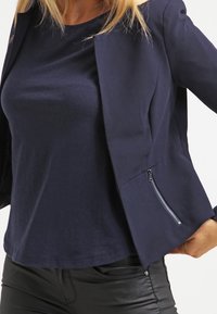 Blazer bleu marine à la coupe ajustée, doté d'une poche zippée sur le côté gauche. Tissu doux, épaules structurées et longueur courte.