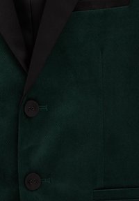 Grön manchesterjacka med svart slag, två svarta knappar och en enda bröstficka. Slät textur med skräddarsydd passform.