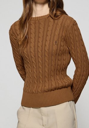 Frau trägt einen braunen Zopfstrickpullover mit geripptem Abschluss und Bündchen, kombiniert mit hellbeigen Hosen, vor einem schlichten Hintergrund.