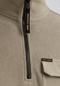 Jersey beige con canalé, de cuello alto, con cierre de media cremallera y cremallera negra. Presenta un bolsillo lateral con una etiqueta "PME LEGEND".