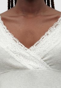 Άτομο με σκούρο δέρμα που φοράει ένα ανοιχτό γκρι ριμπ μπλουζάκι με βαθύ V-λαιμόκοψη διακοσμημένη με λευκά δαντελένια λουλουδένια μοτίβα.