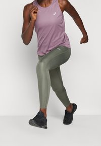 Lilla atletisk tank top med logo, kombineret med grønne leggings med sidelufthuller og sorte løbesko.