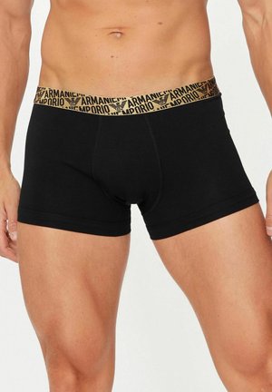 Torso maschile e parte superiore delle cosce indossando boxer aderenti neri con una fascia elastica beige in vita con il marchio Armani Emporio.