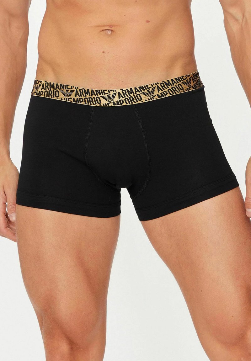 Torso maschile e parte superiore delle cosce indossando boxer aderenti neri con una fascia elastica beige in vita con il marchio Armani Emporio.