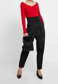 Rotes Langarmoberteil mit einem gewellten Ausschnitt, kombiniert mit hochgeschnittenen schwarzen Hosen, die einen breiten Gürtel haben. Die Accessoires bestehen aus einer kleinen schwarzen Tasche.