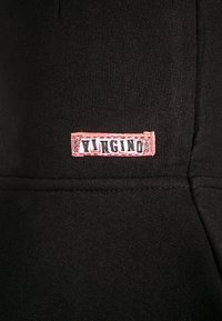 Černá bavlněná tkanina má přišitou růžovou etiketu s textem "YVNGINO" v černé barvě. Hladká textura, minimalistický design bez viditelných vzorů.