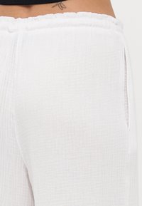 White, textured fabric pants with a loose fit, elastic waistband, and side pockets. The fabric features a soft, crinkled pattern.