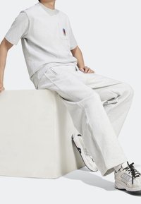 Osoba v světle šedých teplákách, odpovídajícím tričku s malou barevnou nášivkou a bílých teniskách s černými tkaničkami, sedí na bílém bloku.