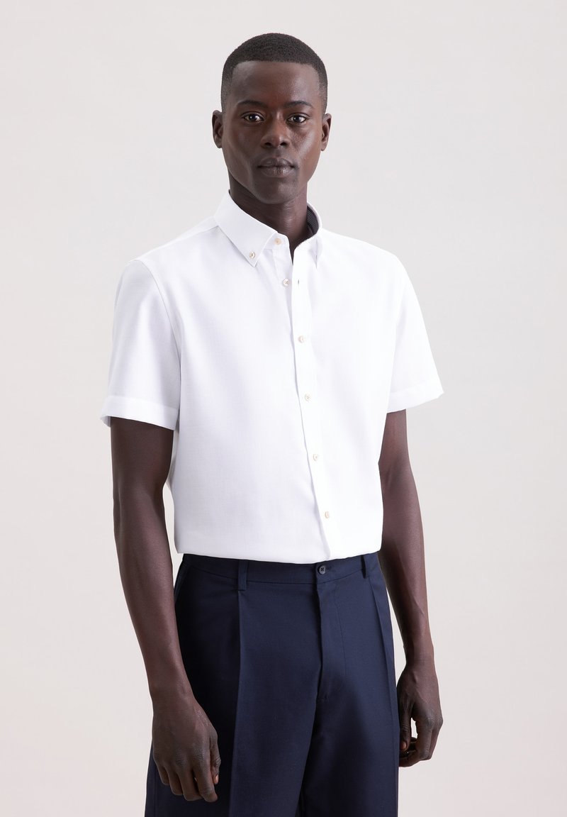 Homme portant une chemise blanche cintrée à manches courtes avec boutons et un pantalon bleu marine foncé, debout devant un fond clair uni.