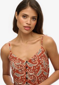 Czerwona sukienka w paisley z cienkimi ramiączkami, zapinana na guziki z przodu, wykonana z teksturowanej tkaniny; posiada wzór wirów i akcenty kwiatowe.