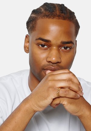 Bague en or sur une main aux doigts détendus, peau lisse, coiffure tressée et portant un T-shirt blanc. Fond neutre.