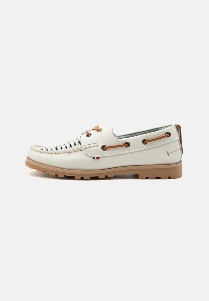 Chaussure bateau en cuir blanc avec devant tissé, lacets marron passés dans des œillets métalliques, semelle en caoutchouc beige, et un petit détail de drapeau tricolore.