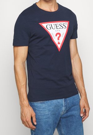 Man wearing a navy blue T-shirt with a red and white triangular Guess logo and light blue denim jeans.