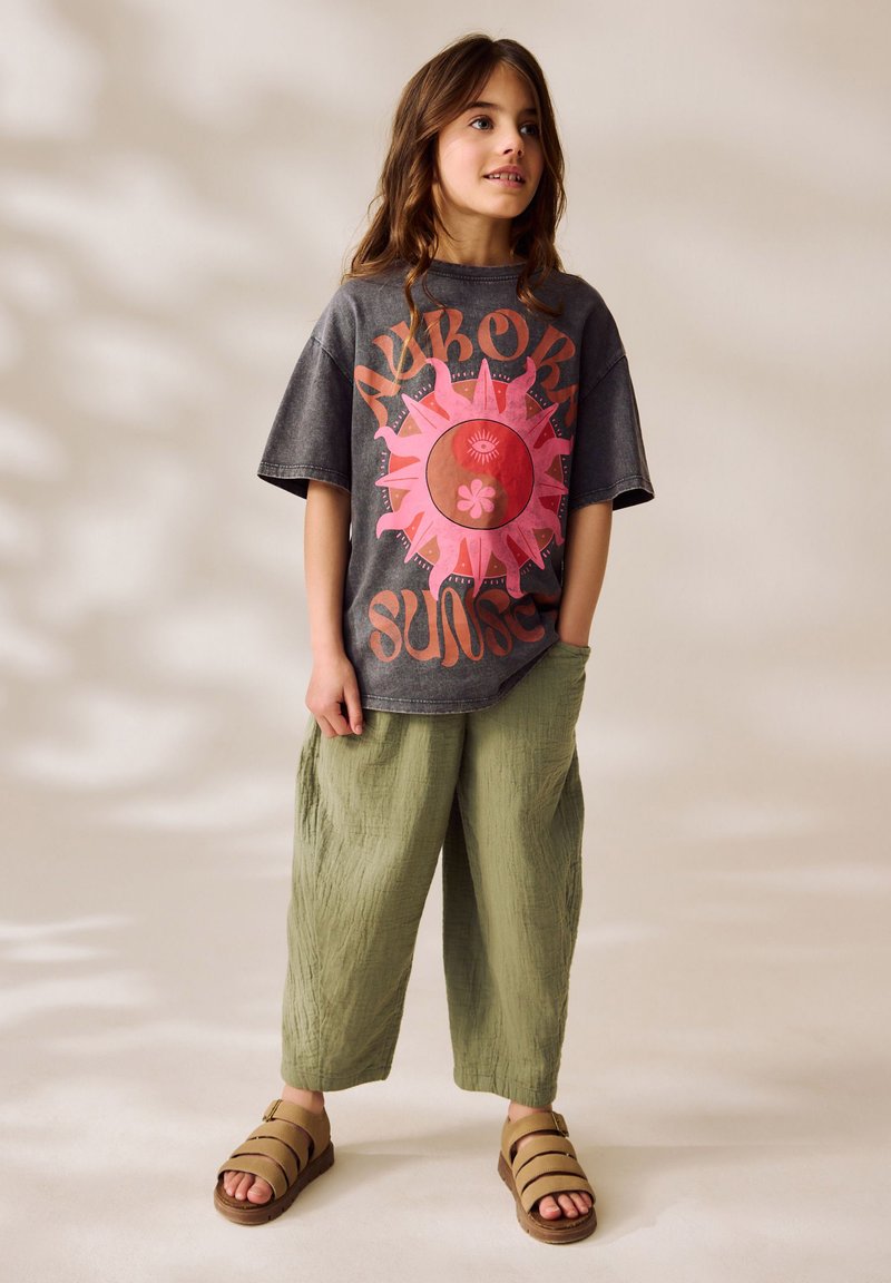 T-shirt graphique gris avec un grand motif de soleil rose, associé à un pantalon en lin vert ample et des sandales en cuir marron à brides. Tenue décontractée et confortable.