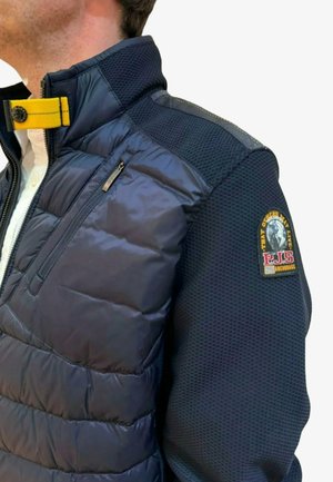 Marineblauwe pufferjas met textuurgebreide mouwen, gele kraagriem, ritssluiting op de borstzak en een ronde badge op de linkerarm.