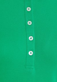 Vihreä neulepolo, jossa on viisi nappia valkoisilla napeilla ja teksturoitu kangas. Lähikuva keskittyy napin yksityiskohtiin.