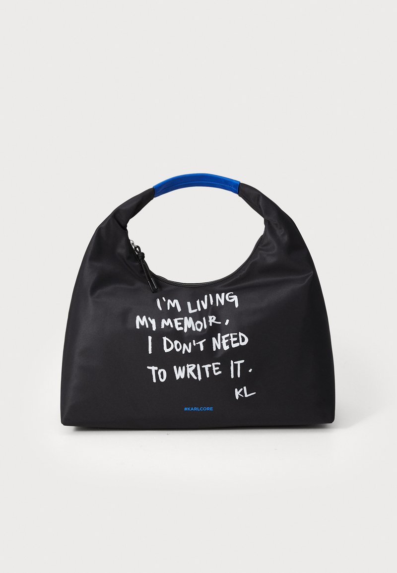 Borsa a tracolla nera con manico blu, con testo bianco che recita "STO VIVENDO LA MIA MEMORIA, NON HO BISOGNO DI SCRIVERLA. KL."