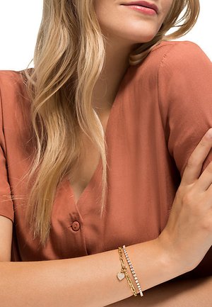 Gold- und Silberarmbänder mit Herzanhänger, geschichtet mit einer kristallbesetzten Kette. Das Modell trägt eine rostfarbene Bluse mit Knopfdetails.