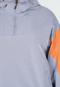 Lys grå pulloverjakke med en orange accent på højre skulder, der har en høj krave, lynlås og et struktureret, let stof.