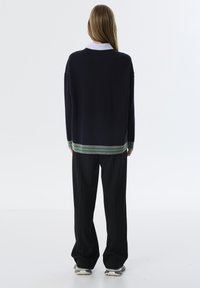 Marineblauwe trui met daaronder een wit met blauwe gestreepte shirt met kraag, voorzien van groene en grijze gestreepte accenten aan de zoom. Brede zwarte broek.
