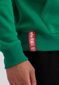Sudadera verde con puños y dobladillo acanalados, con una etiqueta roja que muestra el texto "REMOVE BEFF" en blanco, fabricada en un material de mezcla de algodón.