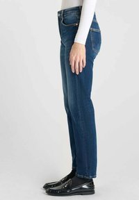 Jean en denim bleu foncé avec une coupe droite, cinq poches et des détails en métal cuivré. Portés avec des mocassins noirs et des chaussettes.