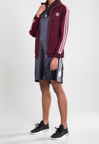 Osoba ubrana w bordową bluzę Adidas na zamek, czarną sukienkę sportową Adidas i czarne trampki z białymi podeszwami, stoi na jednolitym tle.