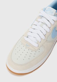 Nike Air Force 1 sneaker featuring a light gray suede upper, white laces, blue accents, perforated toe, and a gum rubber sole.