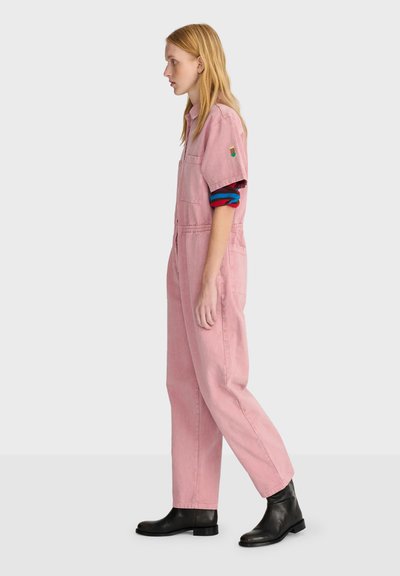 Femme aux cheveux blonds et lisses porte une combinaison rose sur un t-shirt à manches longues rayé et des bottines noires, debout de profil sur fond gris.
