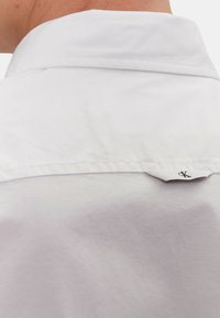 Camicia bianca con colletto strutturato, superficie liscia e una piccola etichetta con logo sul retro. Presenta dettagli di cucitura sottili.