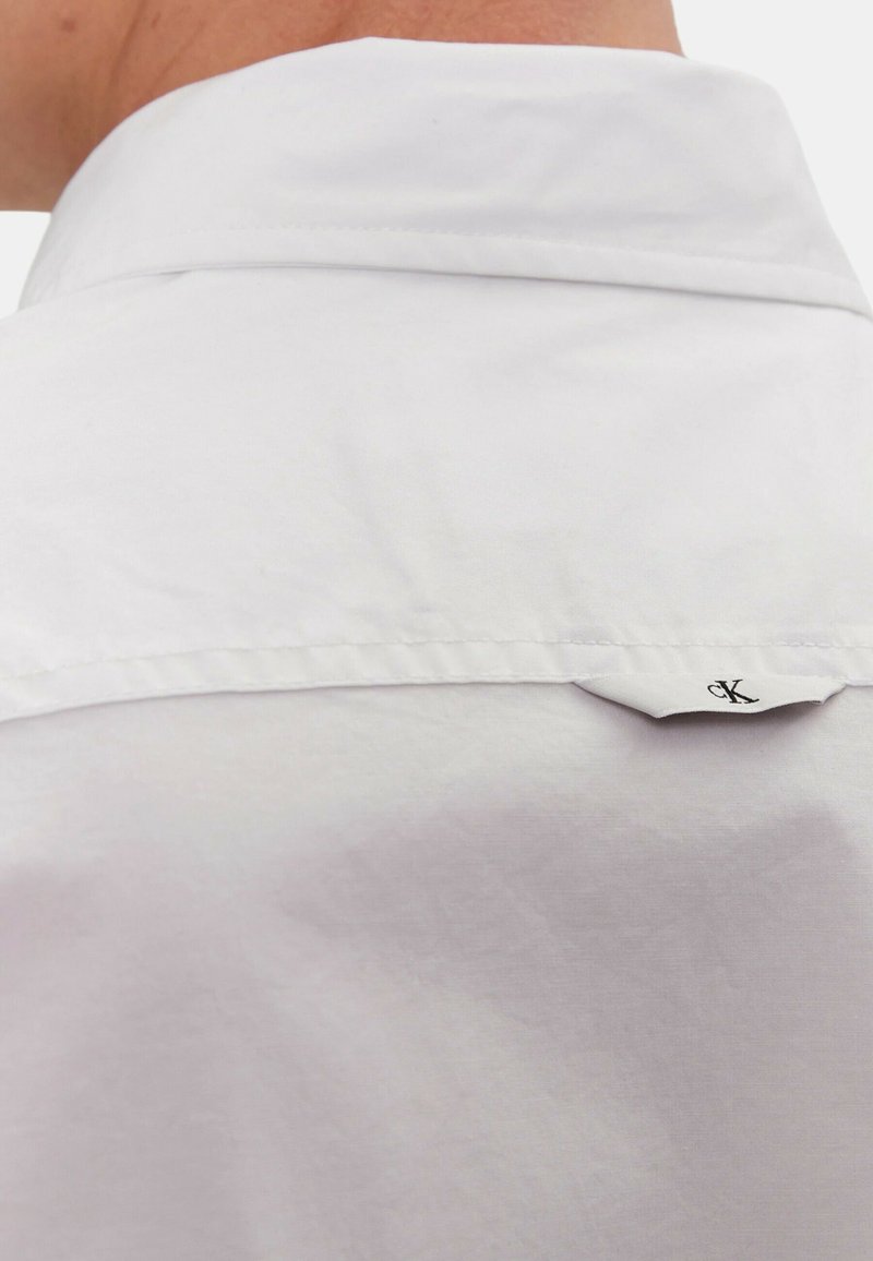 Biała koszula z gładkiego materiału, z wyraźnym kołnierzykiem i małą metką z logo z tyłu. Wyposażona w subtelne detale w postaci przeszyć.