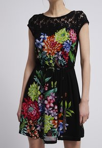 Schwarzes Kleid mit einem Spitzenansatz und bunten Blumenmustern mit Gänseblümchen, Sukkulenten und Schmetterlingen. Enthält einen Bindegürtel in der Taille.