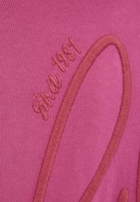 Pink fabric with raised embroidery. Text reads "Since 1981" in light brown, accompanied by an abstract, curved design. Smooth texture.
