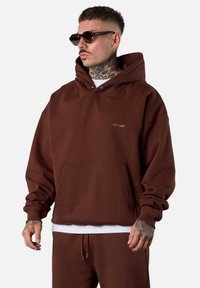 Haut à capuche oversized marron avec une poche avant, arborant un petit logo sur la poitrine et des poignets côtelés. Porté avec un t-shirt blanc en dessous et un pantalon assorti.