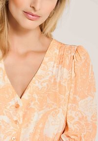 Helles orange Blusenshirt mit Floralprint, V-Ausschnitt, Knöpfen vorne und gepufften, gerafften Ärmeln. Glatte Stoffoberfläche.