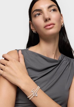 Kobieta o długich ciemnych włosach, ubrana w szary bezrękawnik, nosząca srebrne kolczyki koła oraz warstwowe bransoletki z pereł i srebrnych zawieszek na lewej ręce.