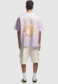 Lilla oversized t-skjorte med sirkulært grafisk trykk med intrikate design på baksiden, kombinert med kremfargede cargo-shortser.