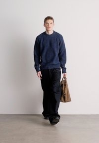 Σκούρο μπλε πλεκτό πουλόβερ, φαρδιά μαύρη παντελόνα και καφέ τσάντα ώμου. Λείες υφές και άνετη εφαρμογή, σε απλό φόντο.