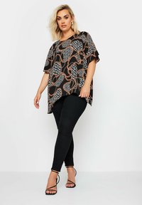 Maglietta nera a maniche corte con un motivo geometrico e catena in arancione e bianco, abbinata a jeans neri skinny e sandali neri con lacci.