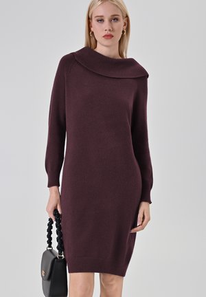Abito lavorato a maglia bordeaux con scollatura ampia e maniche lunghe. Forma aderente, polsini e orlo a costine. Il modello tiene una borsa nera con tracolla intrecciata.