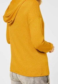 Sweat à capuche jaune moutarde en tissu texturé, avec une grande capuche et des manches longues. Une coupe décontractée avec un ourlet droit et sans embellissements visibles.