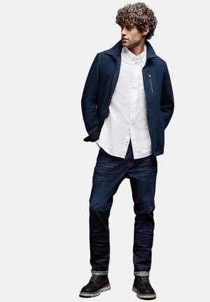 Dunkelblaue Wollmischung-Jacke mit einer Reißverschlusstasche, weißes Hemd mit Knöpfen, dunkle Jeans und schwarze Stiefel mit cremefarbener Sohle.