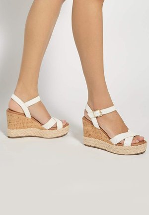 Jambes portant des sandales compensées blanches à bride de cheville, talons en liège et détail en corde tressée sur la semelle, sur un fond uni.