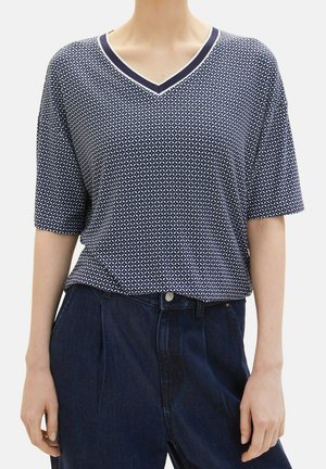 Person wearing a navy blue patterned V-neck short sleeve top tucked into dark blue high-waisted pants.